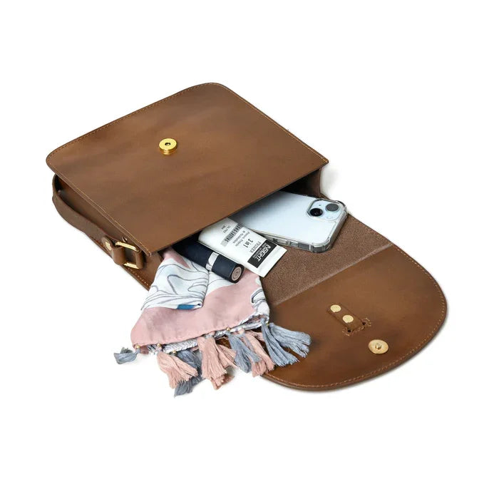 Brown leather designer crossbody bag with magnetic snap, holding a phone, makeup, and a tassel-trimmed scarf.
