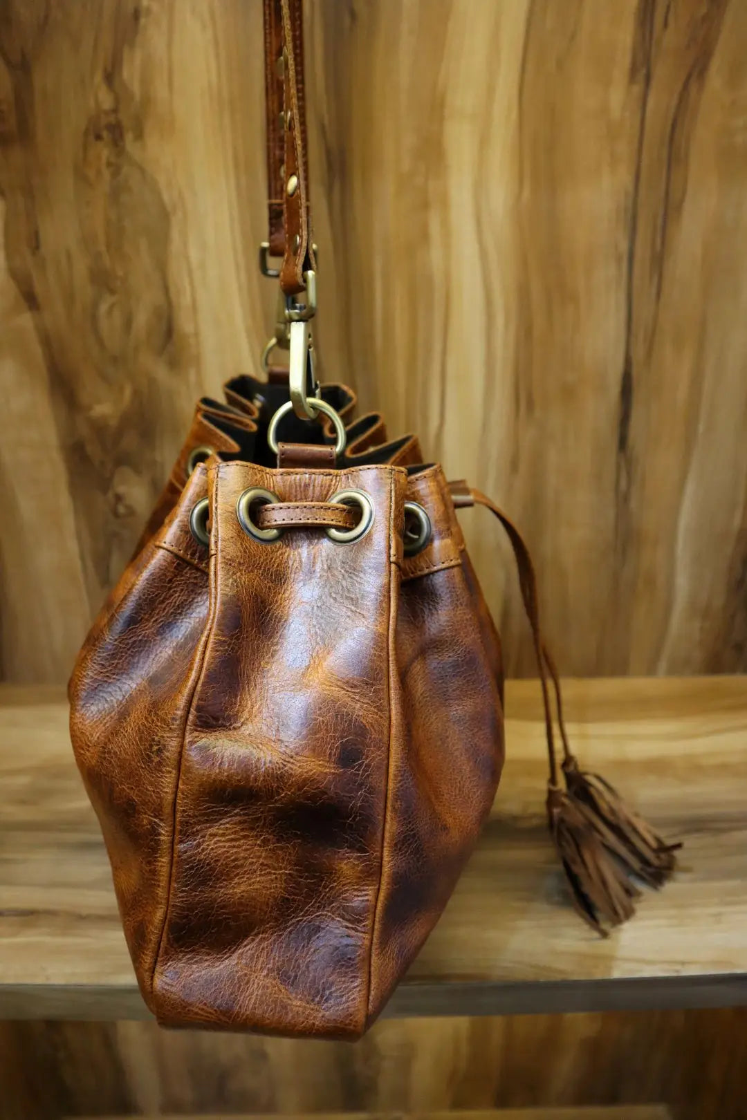 Distressed brown leather bucket bag with brass grommets, adjustable shoulder strap, and tassel detail. Trunk-style.