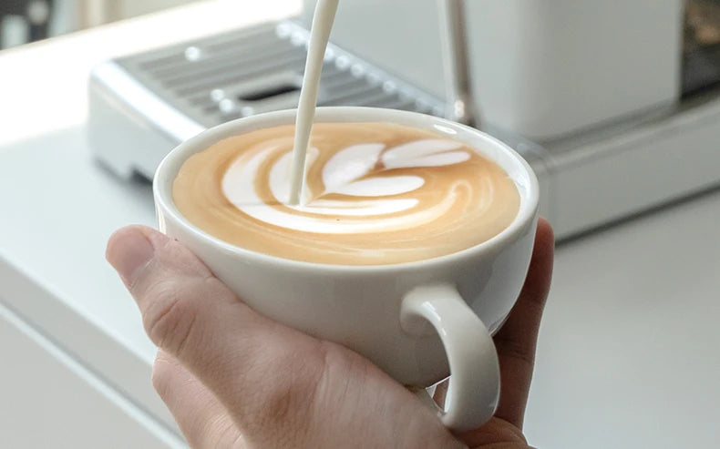 White espresso machine pouring steamed milk into a ceramic latte cup with leaf latte art.