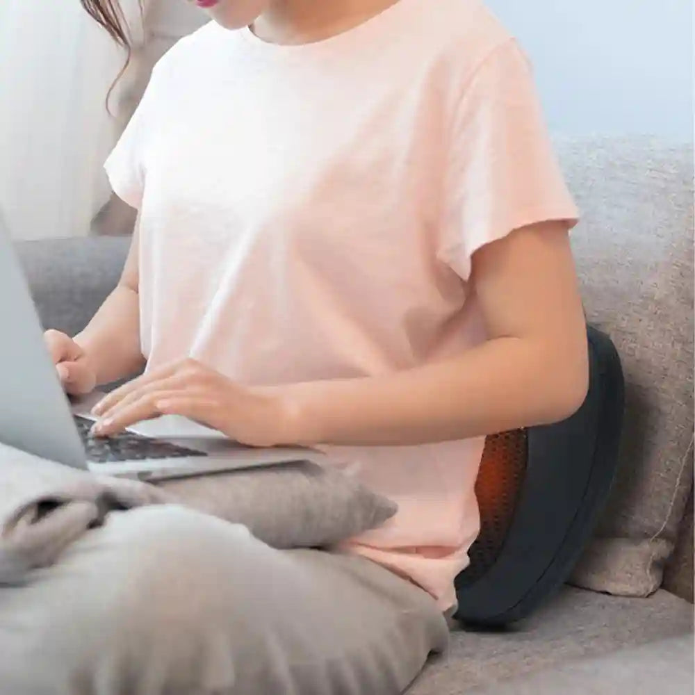 A woman in a pink shirt uses a black, round, mesh Shiatsu massage pillow behind her back while working on a laptop.