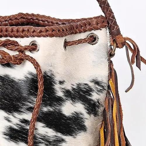 Vintage black and white cowhide bucket bag with braided brown leather trim, drawstring closure, and side fringe.