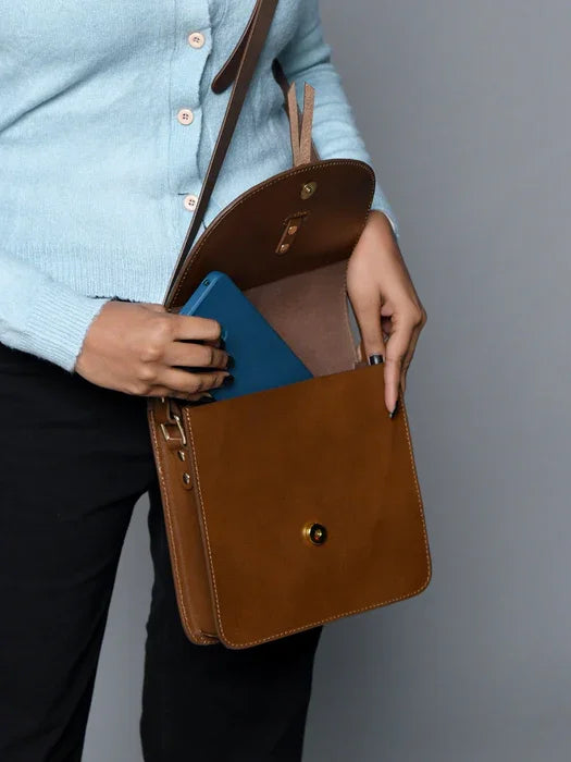 Luxury brown leather designer crossbody bag with gold snap closure, worn with a light blue sweater.