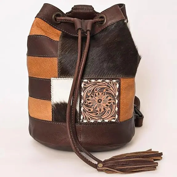 Brown leather bucket bag with western floral tooling, patchwork hair-on hide, drawstring closure, and tassel detail.