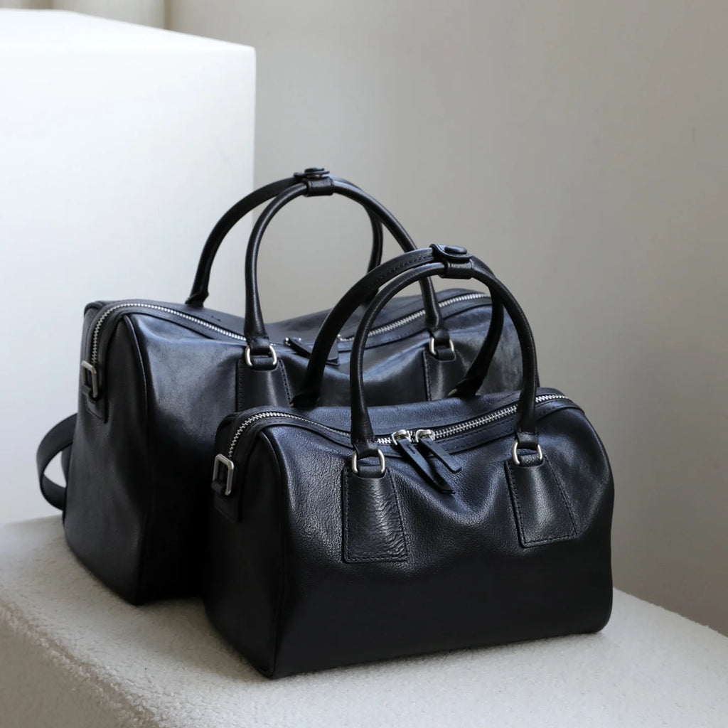 Two black vegetable-tanned leather designer Boston bags with minimalist design, dual zippers, and sturdy top handles.