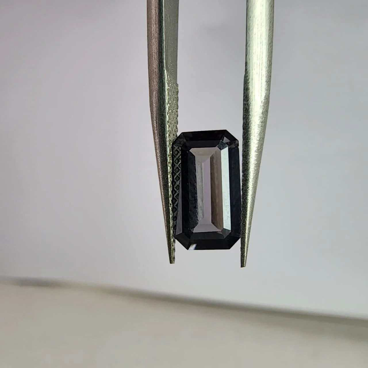 Dark gray spinel gemstone, emerald-cut, held between metal tweezers