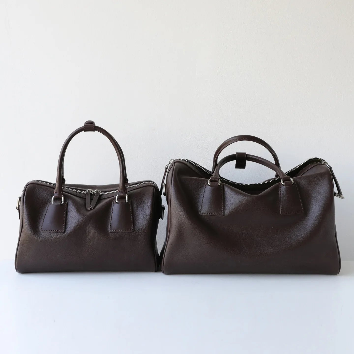 Two dark brown vegetable-tanned leather Boston tote bags with minimalist design, silver zippers, and durable handles.