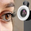 Black clip-on macro lens for smartphones with a circular LED ring light attached, positioned near a human eye.