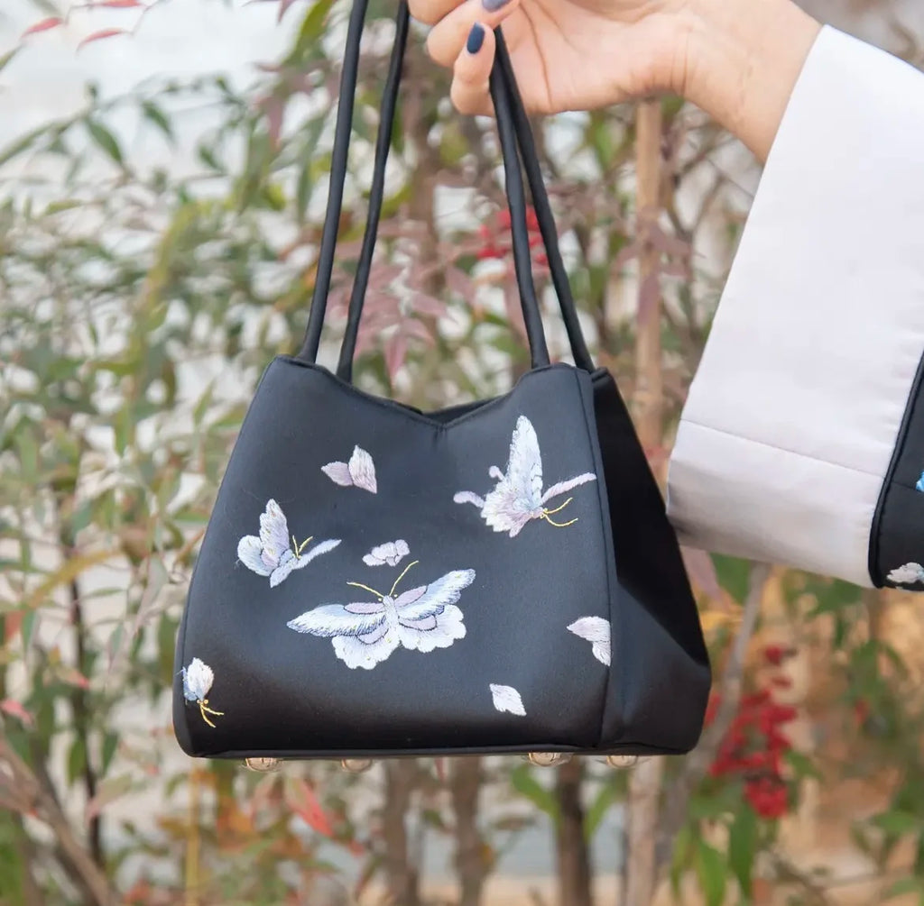Black satin shoulder bag featuring delicate white and purple butterfly Su embroidery and thin top handles.