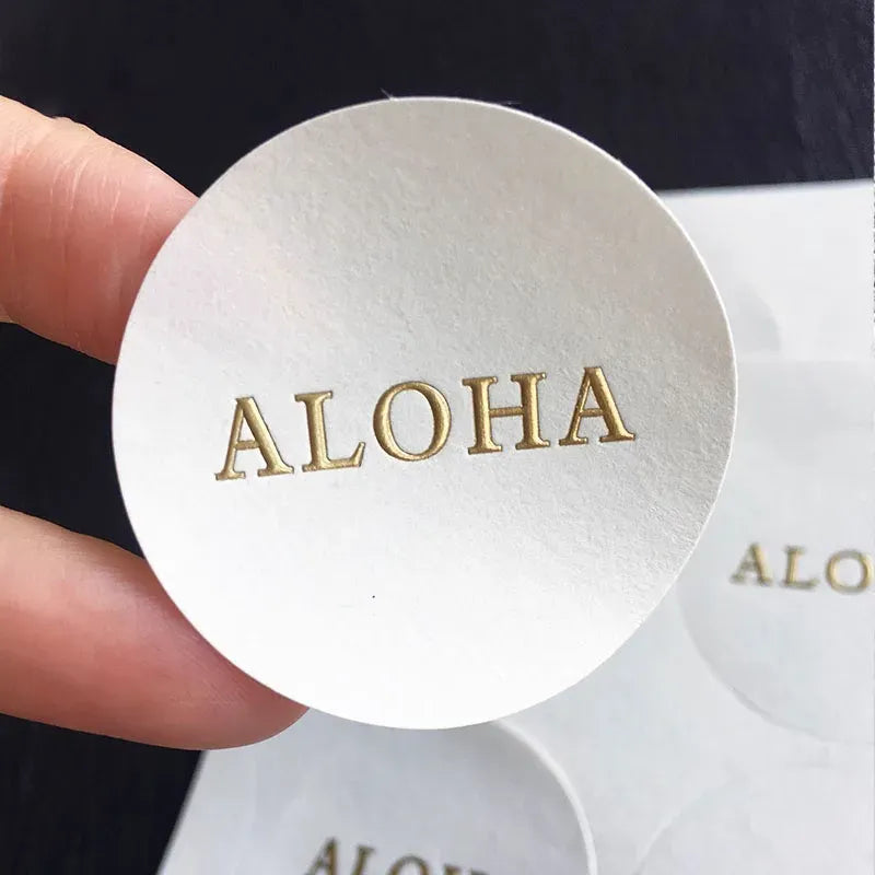 White round sticker label with gold foil ALOHA logo, embossed.
