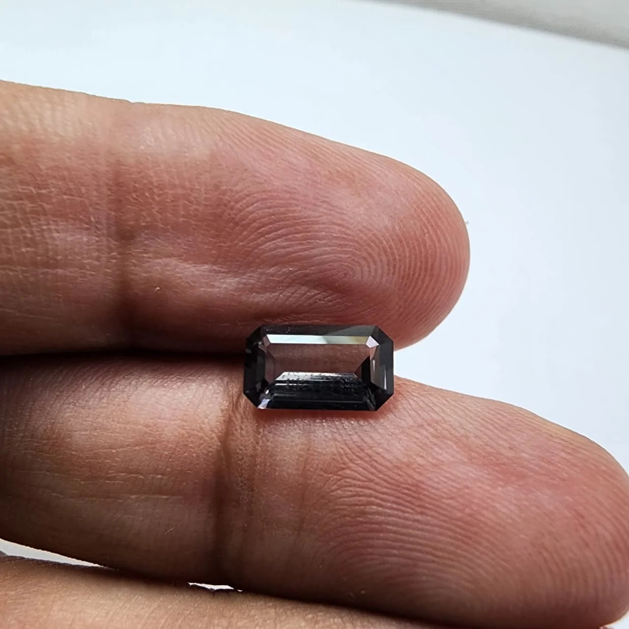 Dark gray emerald-cut spinel gemstone on fingertip, crisp step facets