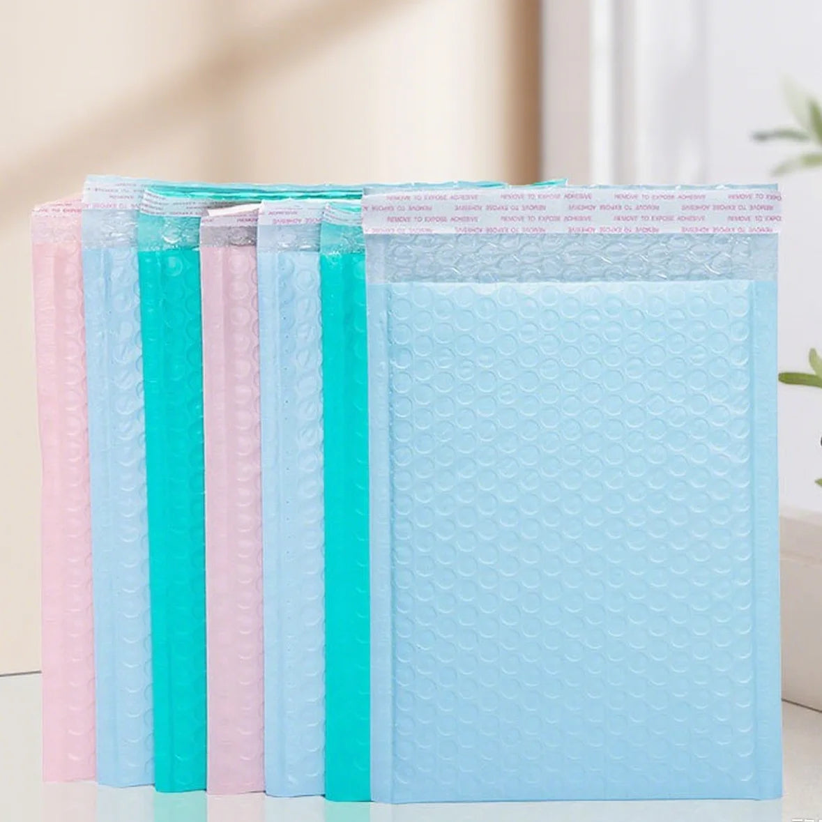 Multi-color bubble mailer bags stack: light blue, teal, and pink with self-seal flap