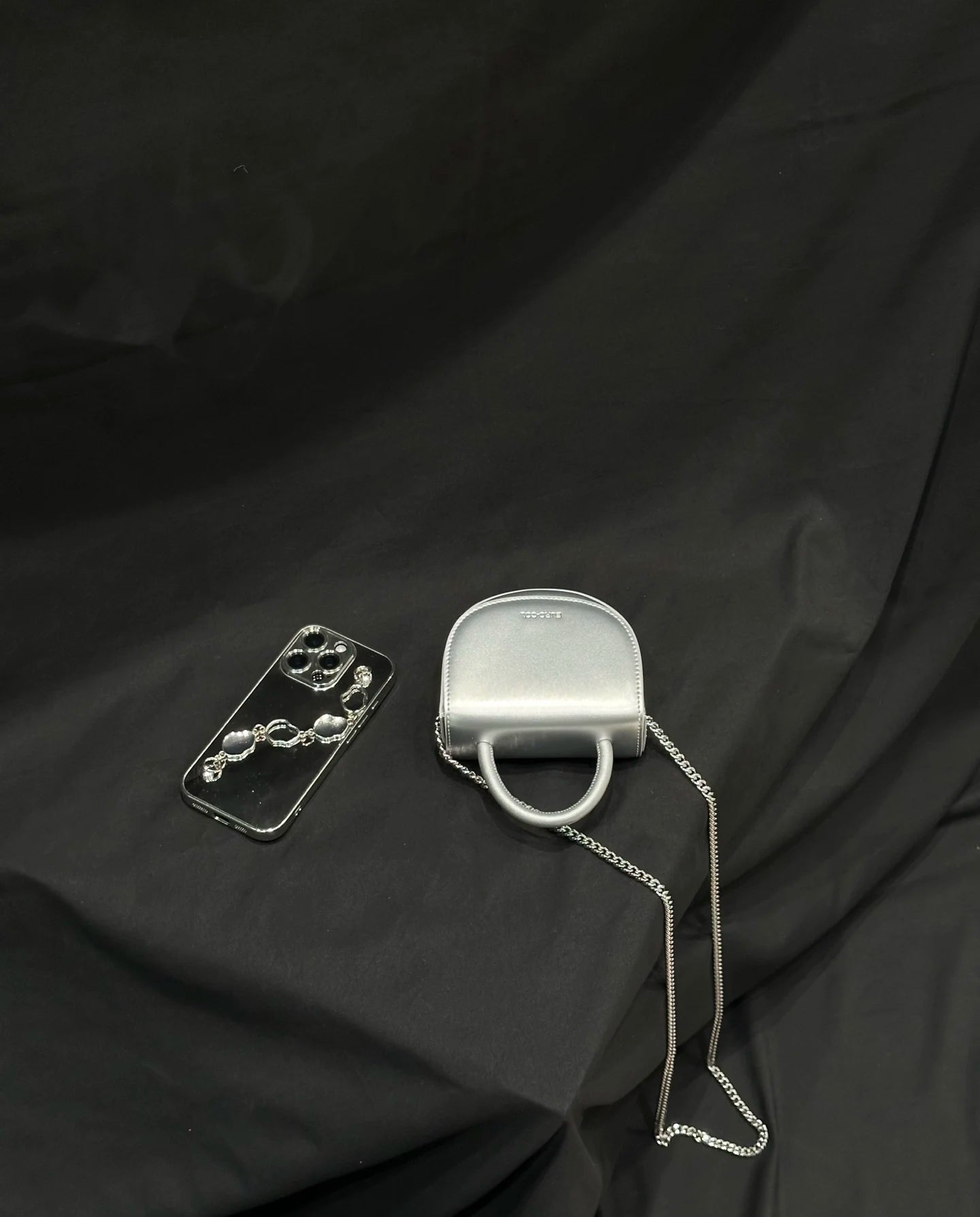 Metallic silver cowhide leather mini evening clutch with top handle, chain strap, and designer style on black fabric.