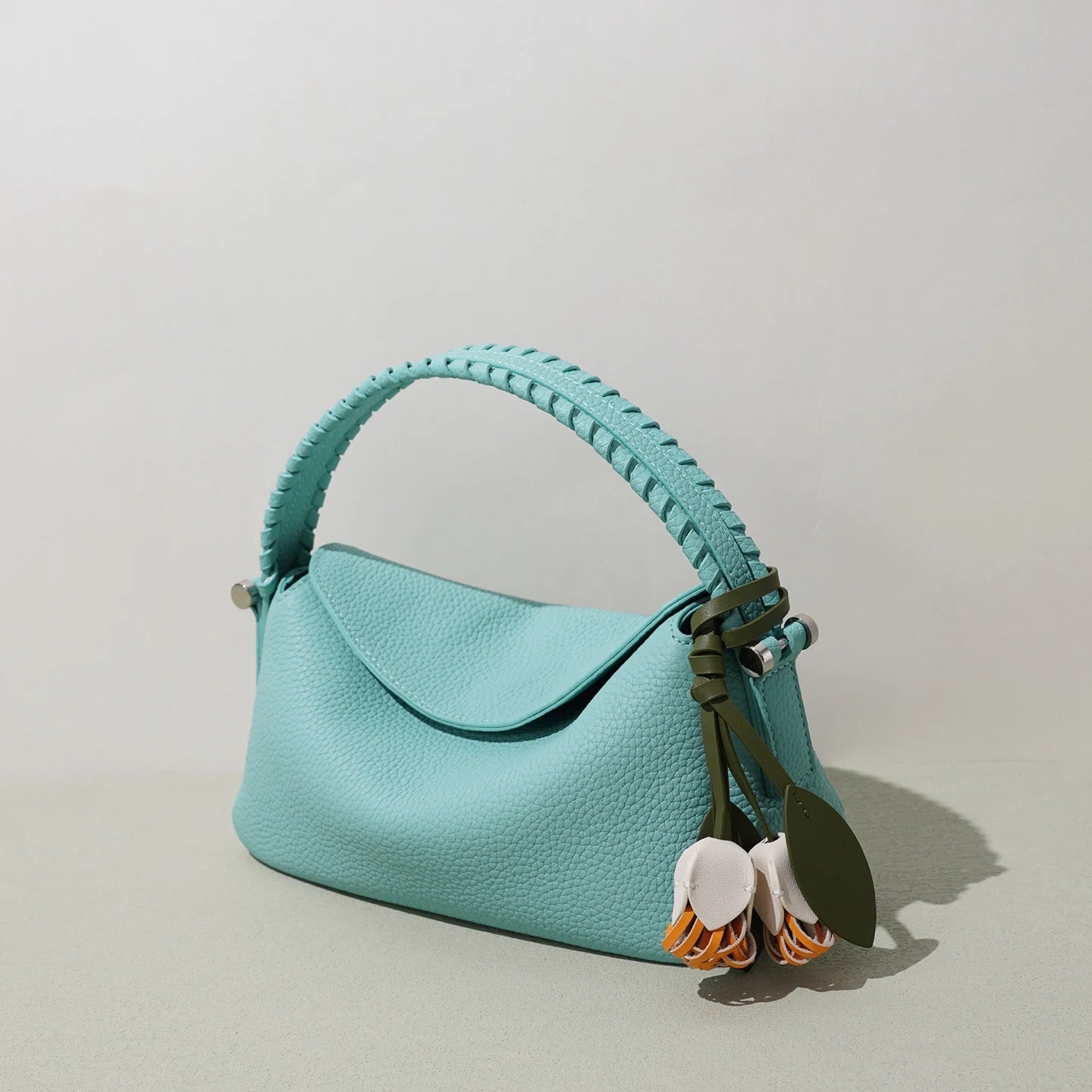 Small turquoise genuine leather designer crossbody bag with a braided handle and decorative floral charm.