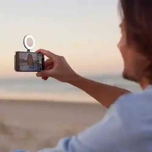 Small circular white LED ring light clipped to a smartphone for illuminating selfies at the beach.