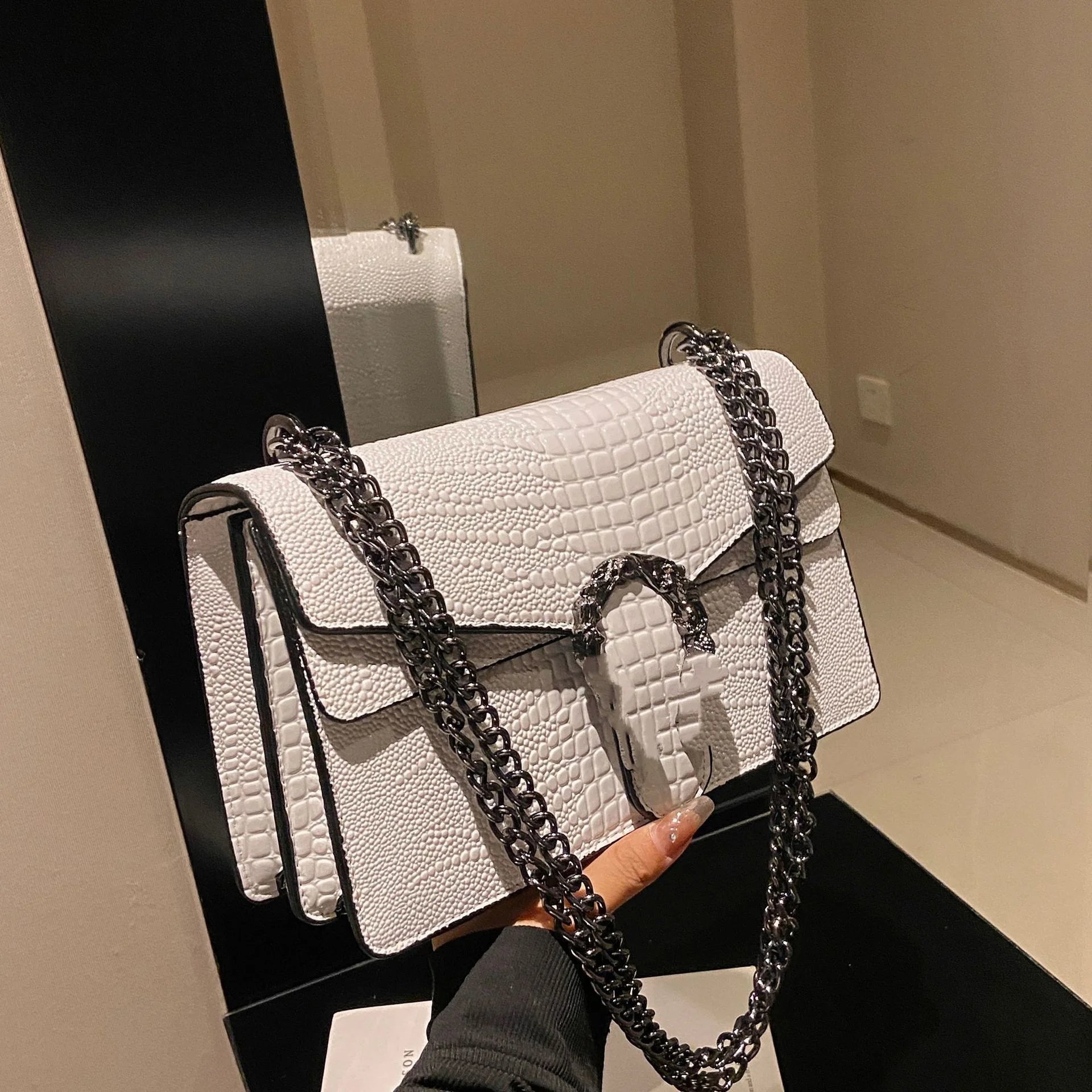 White faux-crocodile crossbody trunk bag with dark metallic chain strap and signature metal clasp closure.