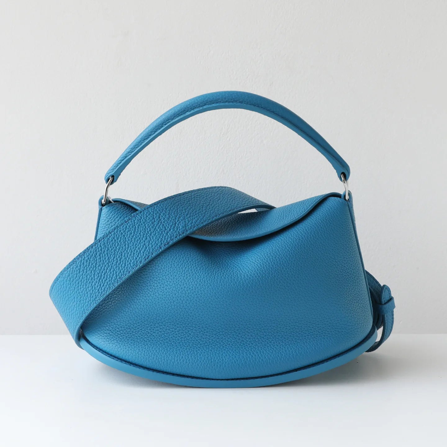 Blue designer handbag in pebbled cowhide leather with a pillow-shaped silhouette, top handle, and magnetic flap.