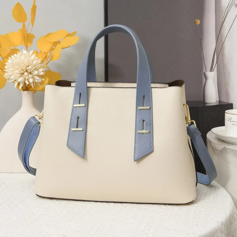 Cream designer handbag with blue leather trim, gold metal accents, structured silhouette, and detachable shoulder strap.