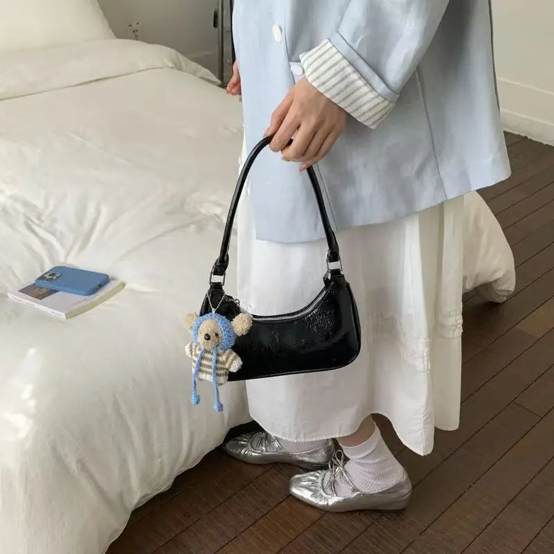 Black patent leather dumpling-style shoulder bag with teddy bear charm and zipper closure.