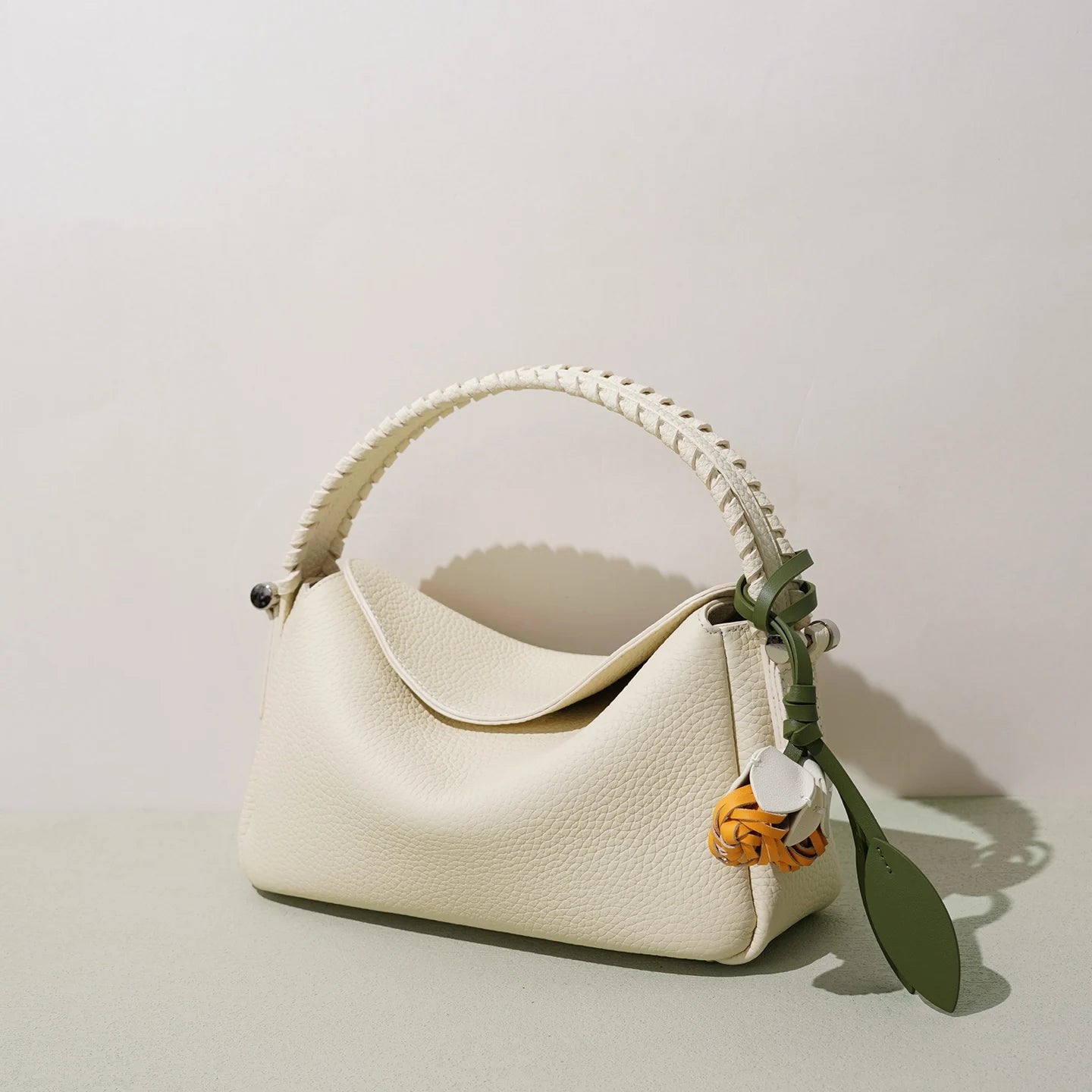 Cream genuine leather designer handbag with woven handle, small tote silhouette, and decorative flower charm.