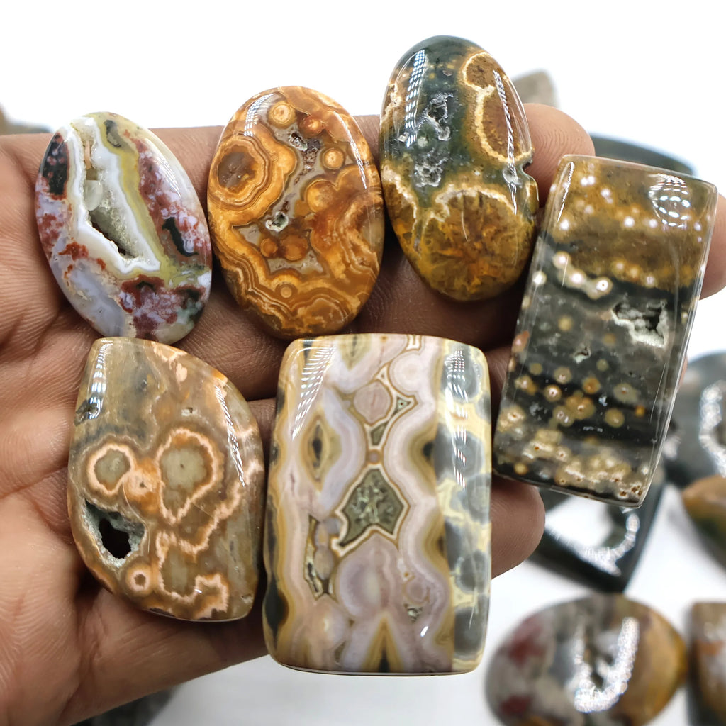 Hand holding ocean jasper cabochons, mixed shapes with white, brown, and gold banded patterns