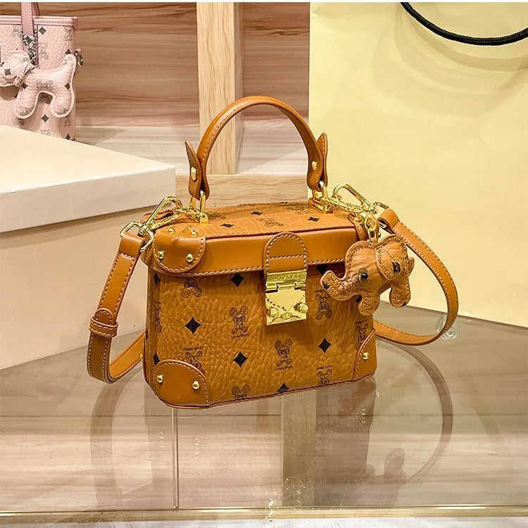 Brown cowhide trunk handbag with logo print, gold hardware, top handle, shoulder strap, and elephant charm.