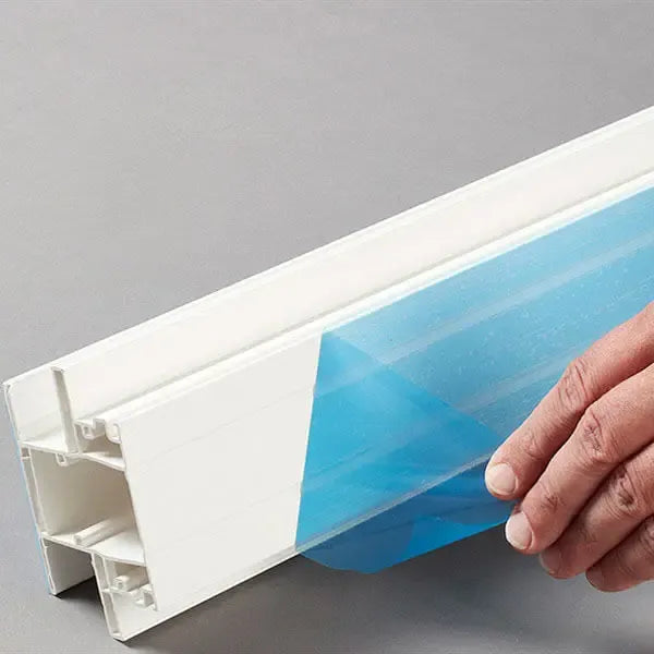 Hand pressing transparent plastic film onto white window profile with protective tape