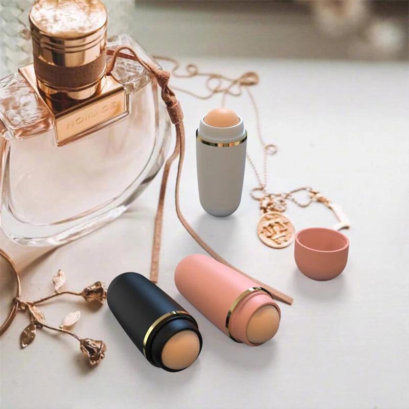 Three volcanic stone oil-absorbing rollers in matte black, pink, and grey, arranged near perfume and gold jewelry.