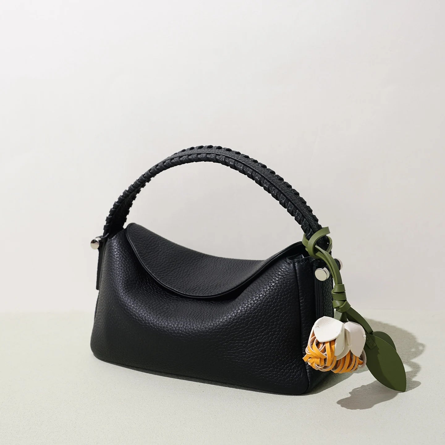Black pebbled leather designer handbag with woven handle, shoulder strap, and cute flower charm.