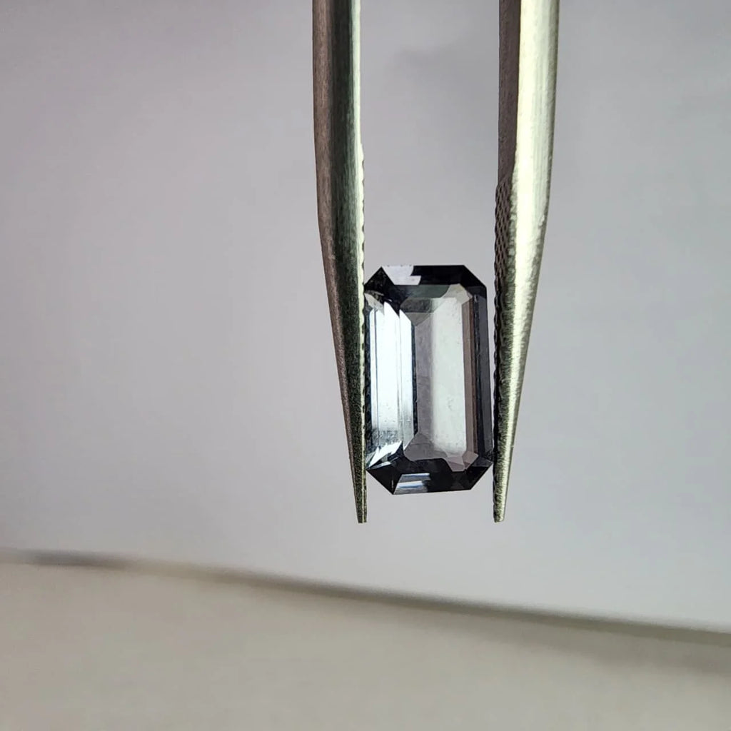 Dark gray spinel gemstone, emerald-cut, view from the side with tweezers