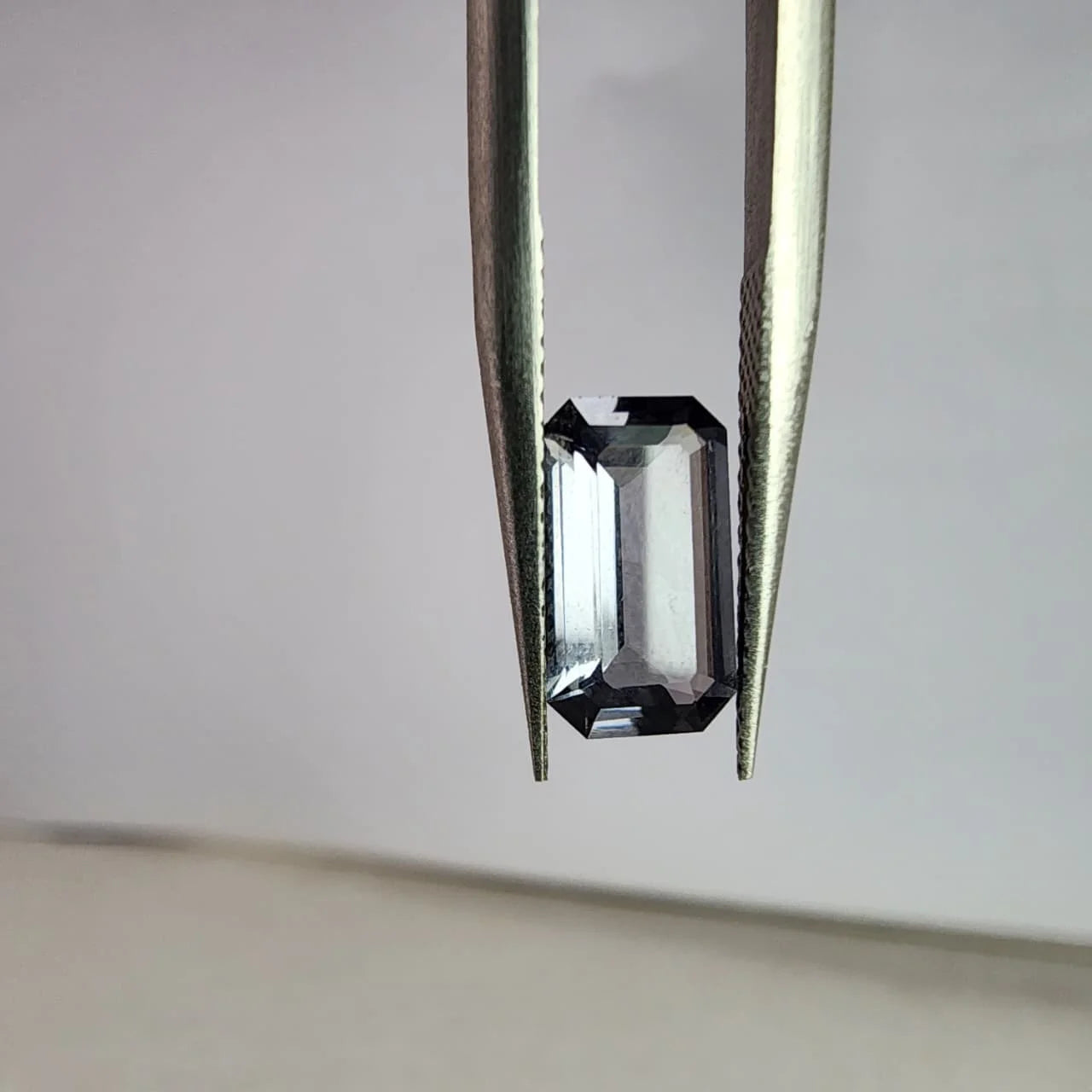 Dark gray spinel gemstone, emerald-cut, view from the side with tweezers