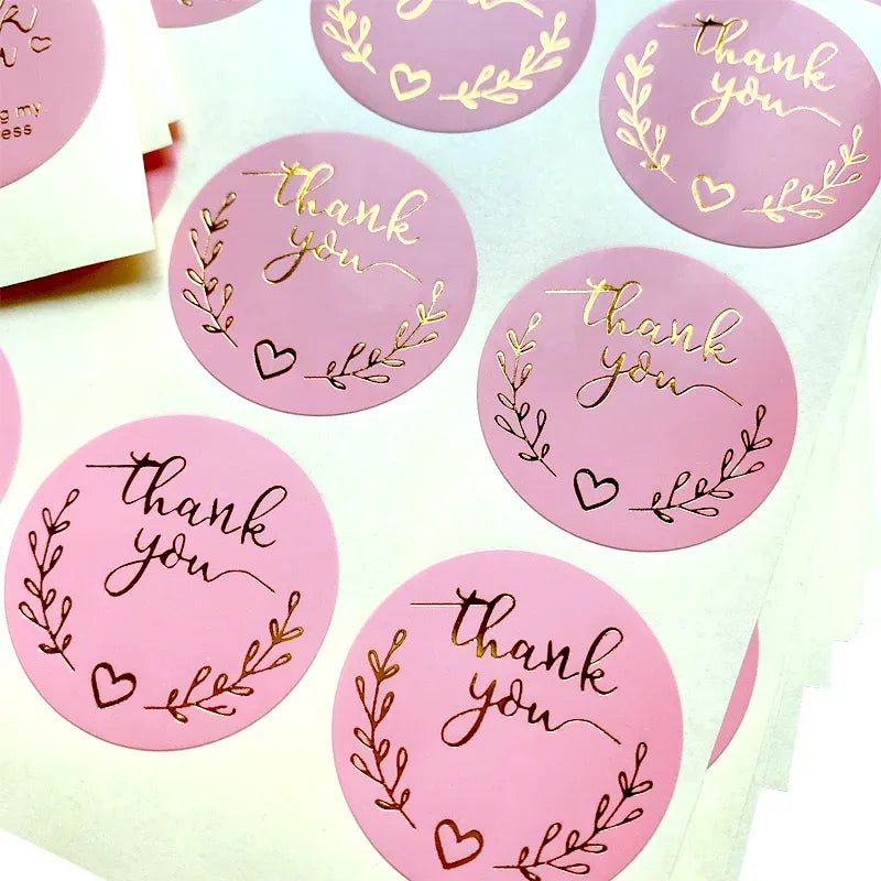 Pink round thank you stickers with gold-foil script, wreath branches, and hearts