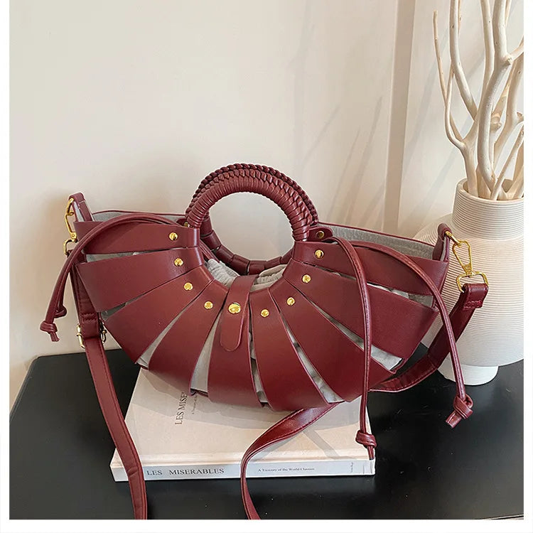 Maroon half-moon crossbody bag with hollow-out leather panels, round braided handle, and gold studs. Casual luxury.