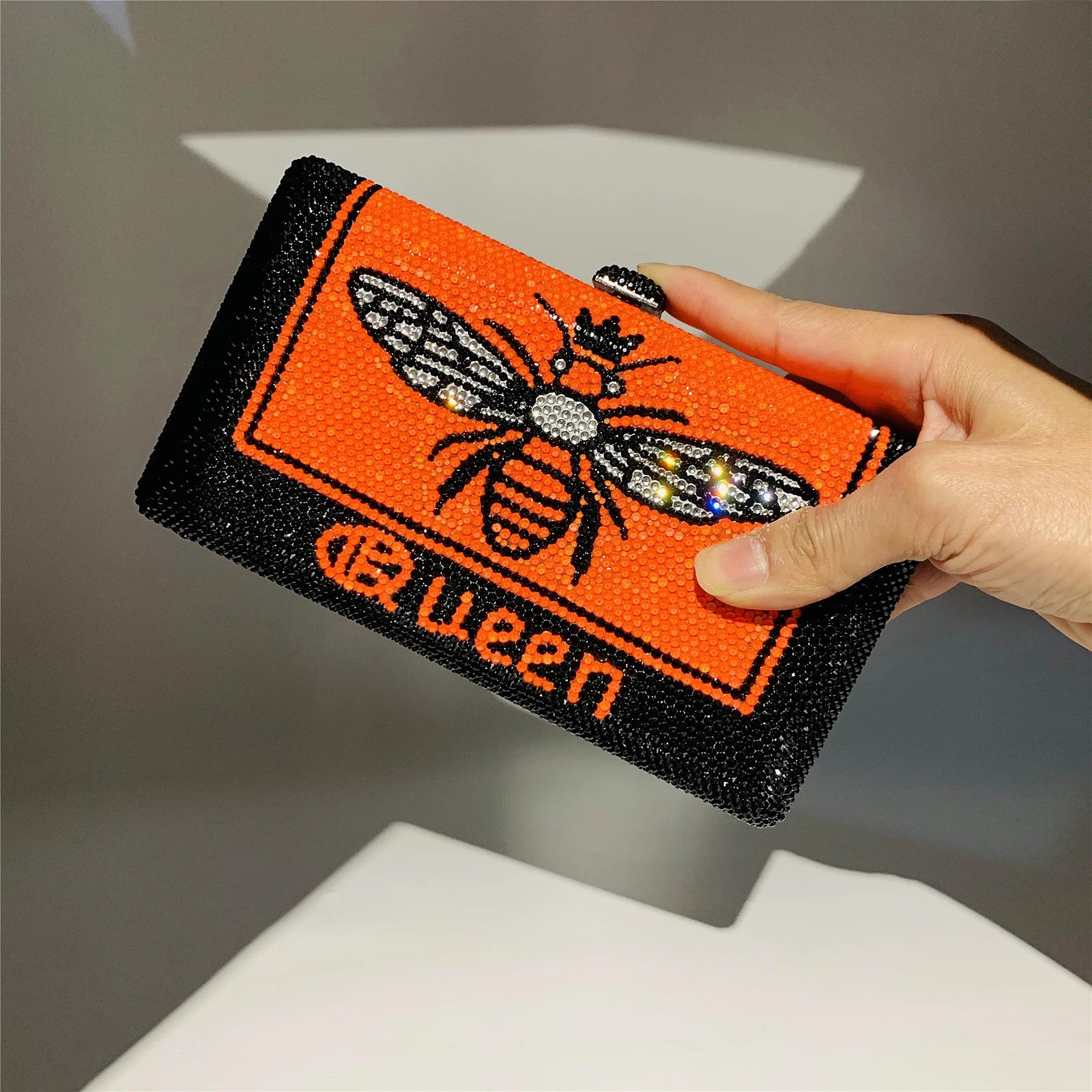 Orange and black rhinestone hard shell evening clutch with "Queen" text and a jeweled bee design. Designer handbag.