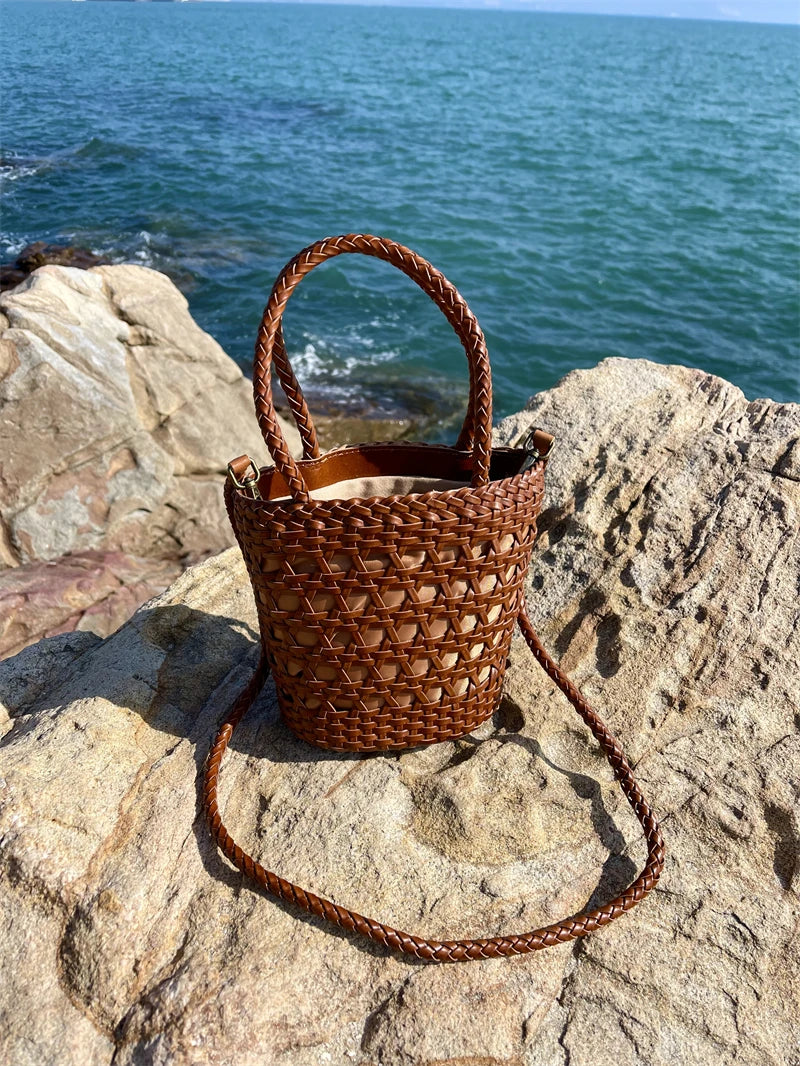 Brown handwoven leather bucket bag with cross-weave cannage design, top handles, and shoulder strap on a seaside rock.