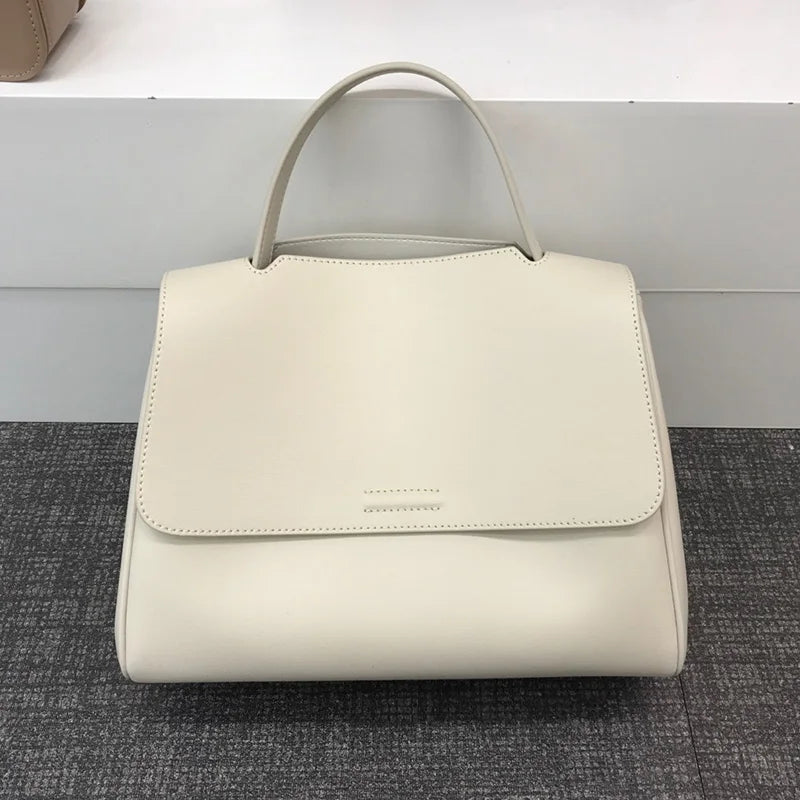 Cream minimalist designer handbag in smooth leather with a structured flap, top handle, and simple stitched detail.