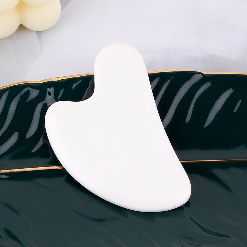 White, heart-shaped resin gua sha massage tool resting on a dark green leaf-shaped decorative plate.