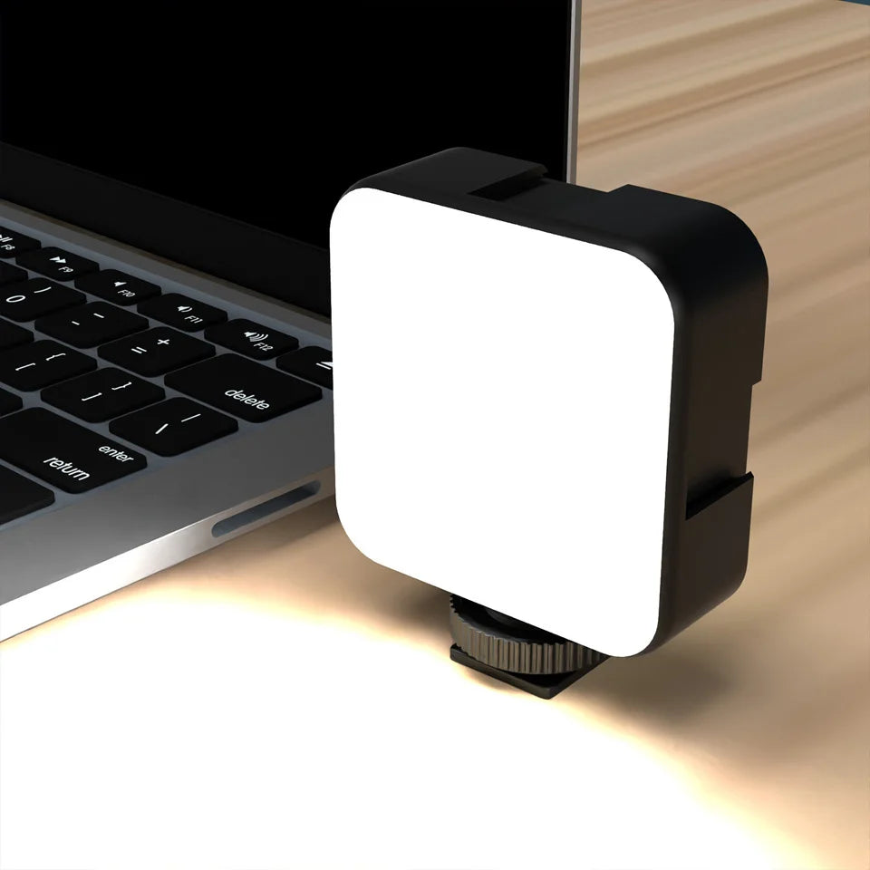 Small square LED video light with a black frame and white diffuser, mounted on a base next to a laptop for lighting.