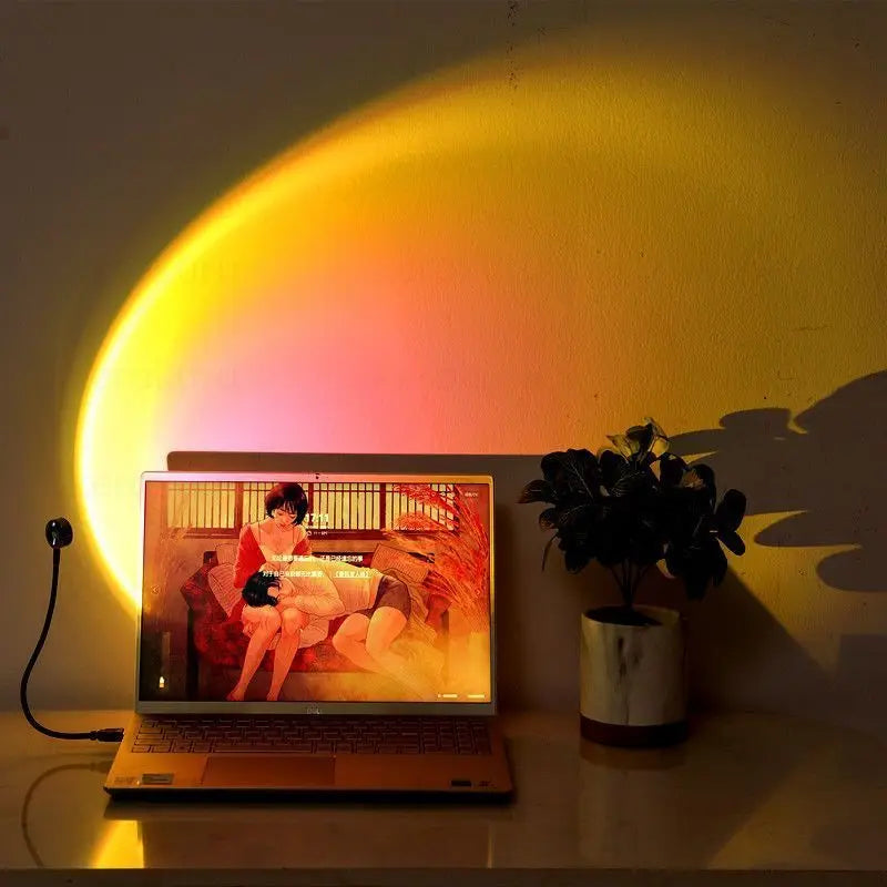 Small USB sunset lamp projecting a yellow and orange arc on a wall behind a laptop and a small potted plant.