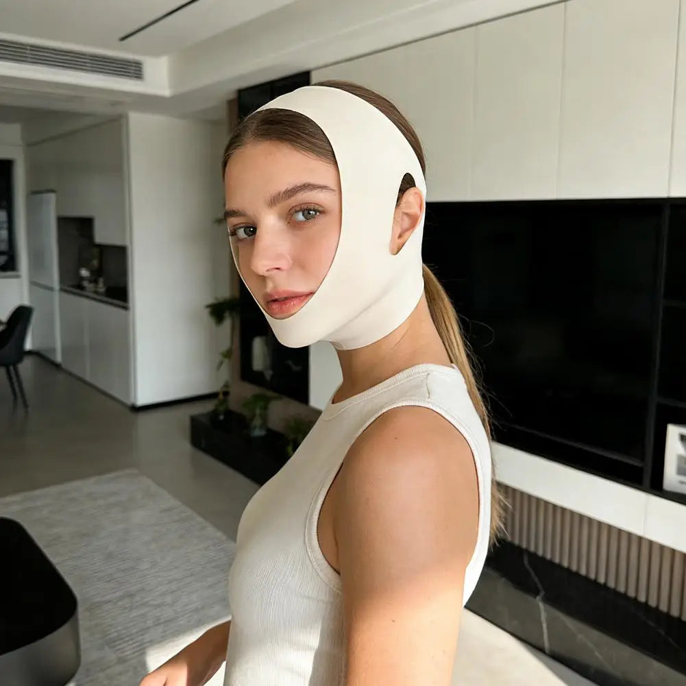 A woman wearing a beige, smooth, elastic V-face shaper belt around her chin and head in a modern living room.