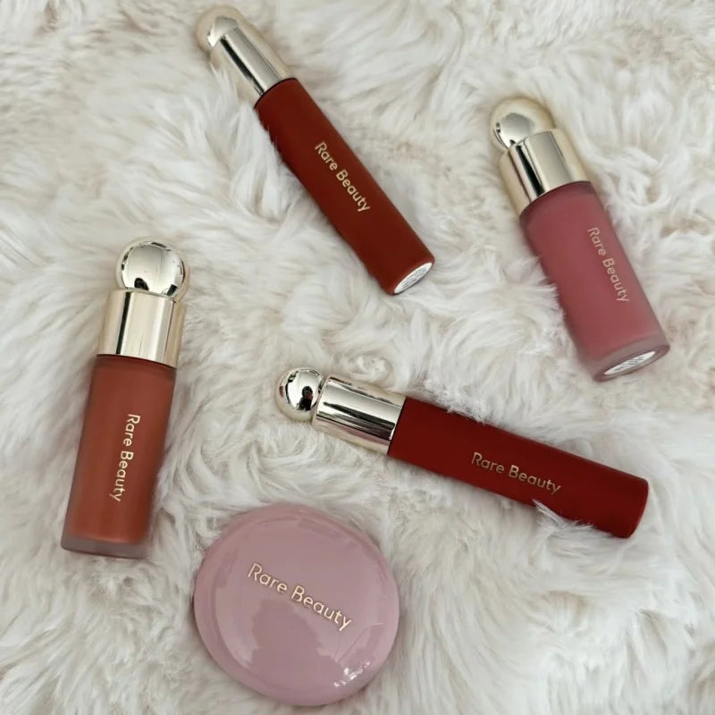 Rare Beauty liquid blush, highlighter, and lip makeup set in assorted matte and dewy shades on a white faux fur rug.