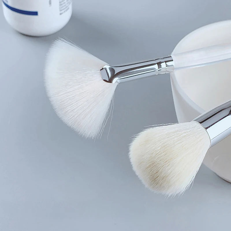 Soft white facial mask fan brush and rounded skin care cleansing brush with silver handles on gray background.