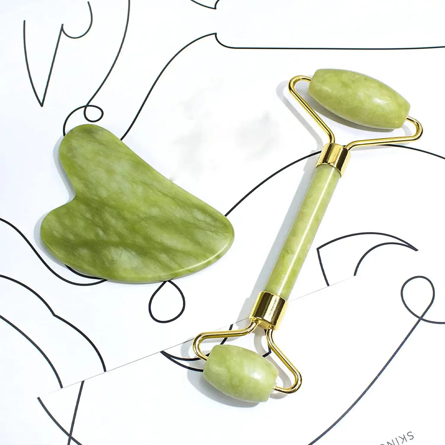 Green jade facial roller and heart-shaped gua sha scraping tool with gold metal accents on a white patterned background.