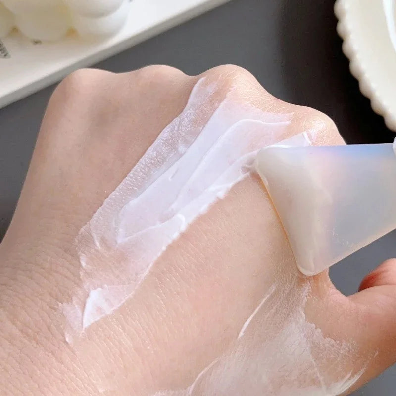A translucent, flexible silicone spatula spreads white facial mask cream smoothly across the back of a hand.