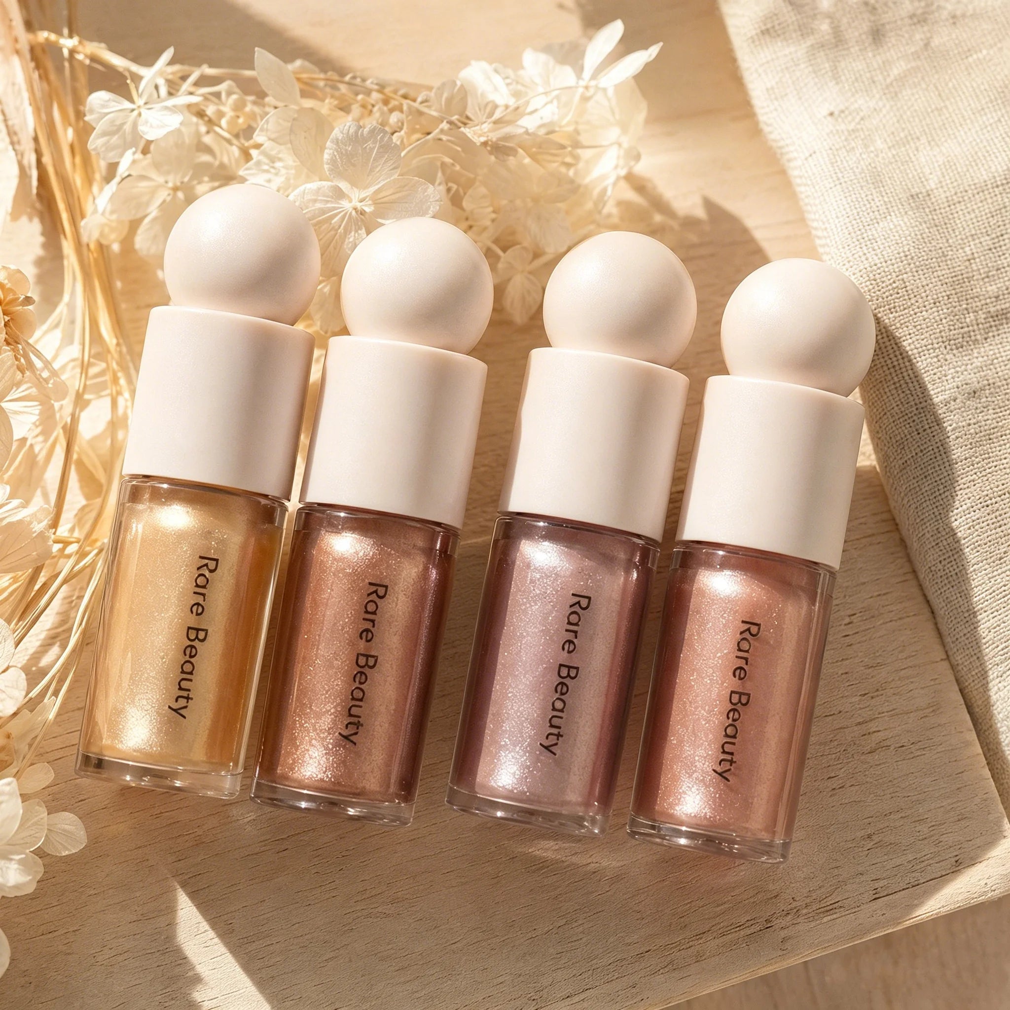 Four Rare Beauty liquid highlighters in shimmer shades with rounded white caps on a wooden surface with white flowers.