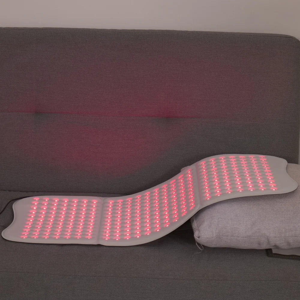 A flexible, light grey red light therapy mat with numerous glowing red LED lights laid out on a grey sofa.