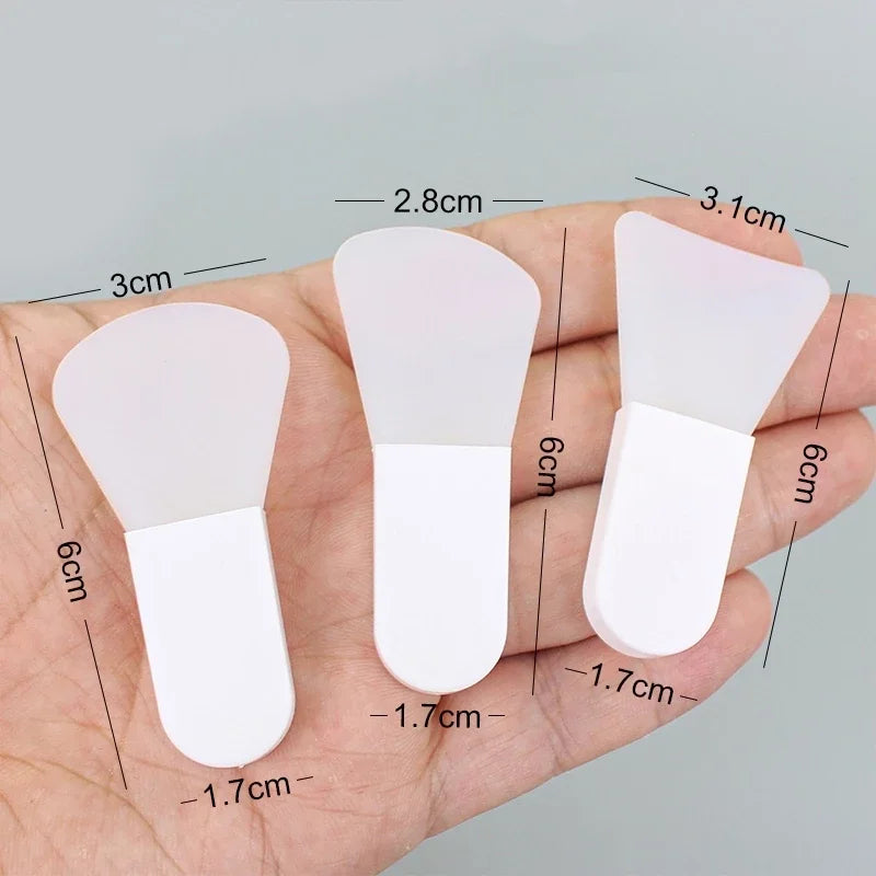 Three white silicone facial mask spatulas of varying head shapes held in a palm, with size dimensions labeled.