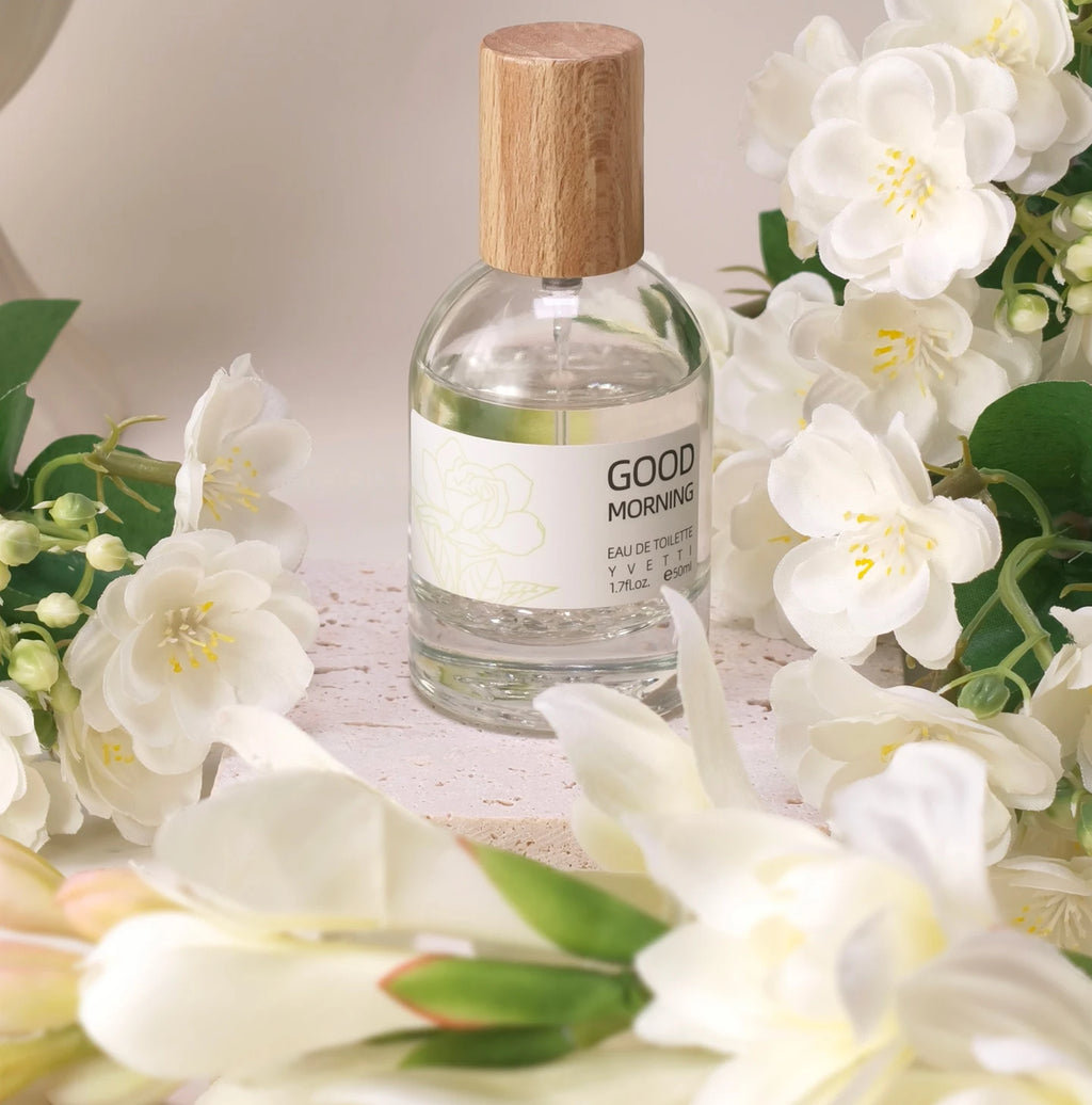 Clear glass perfume bottle with wooden cap, Good Morning label, surrounded by elegant white gardenia flowers.