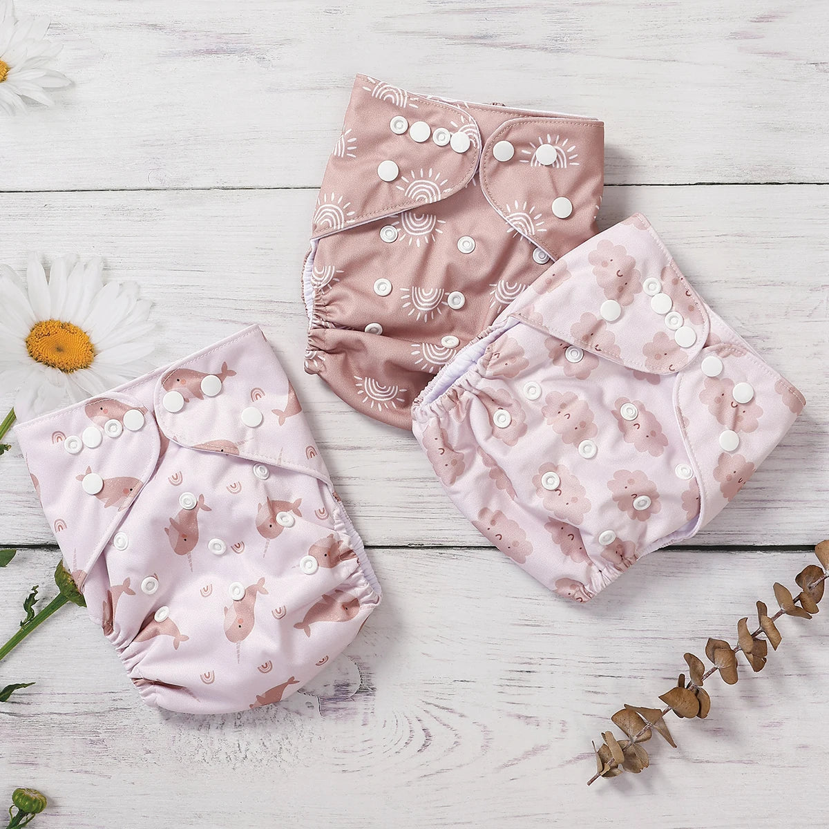 Three reusable cloth baby diapers featuring cute sun, narwhal, and cloud prints with adjustable snap closures.