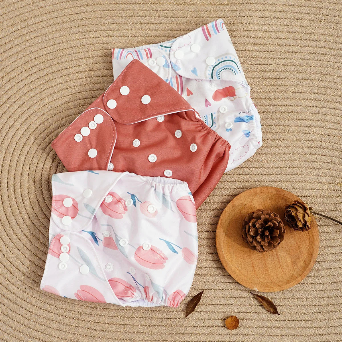 Three adjustable cloth baby diapers in rainbow, solid terracotta, and floral tulip prints on a textured surface.