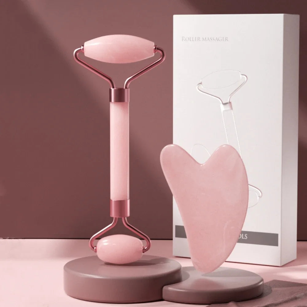 Pink resin dual-ended facial roller and heart-shaped gua sha board with a white product box on display stands.